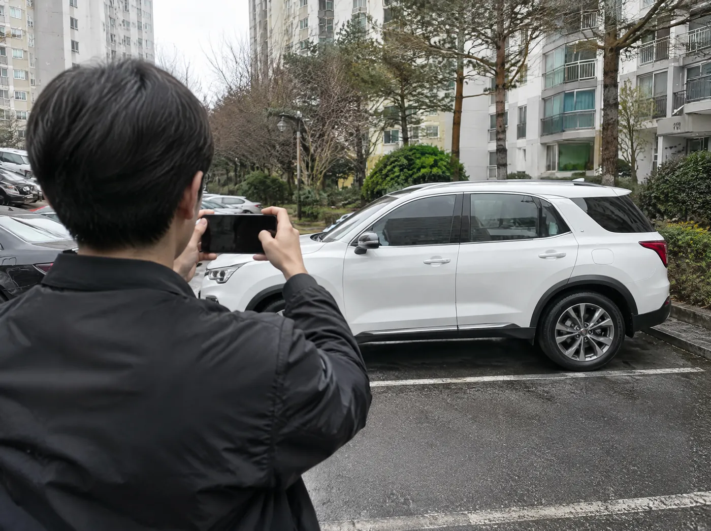 차 주인이 주차장에서 스마트폰으로 자기 SUV 옆모습을 찍는 뒷모습